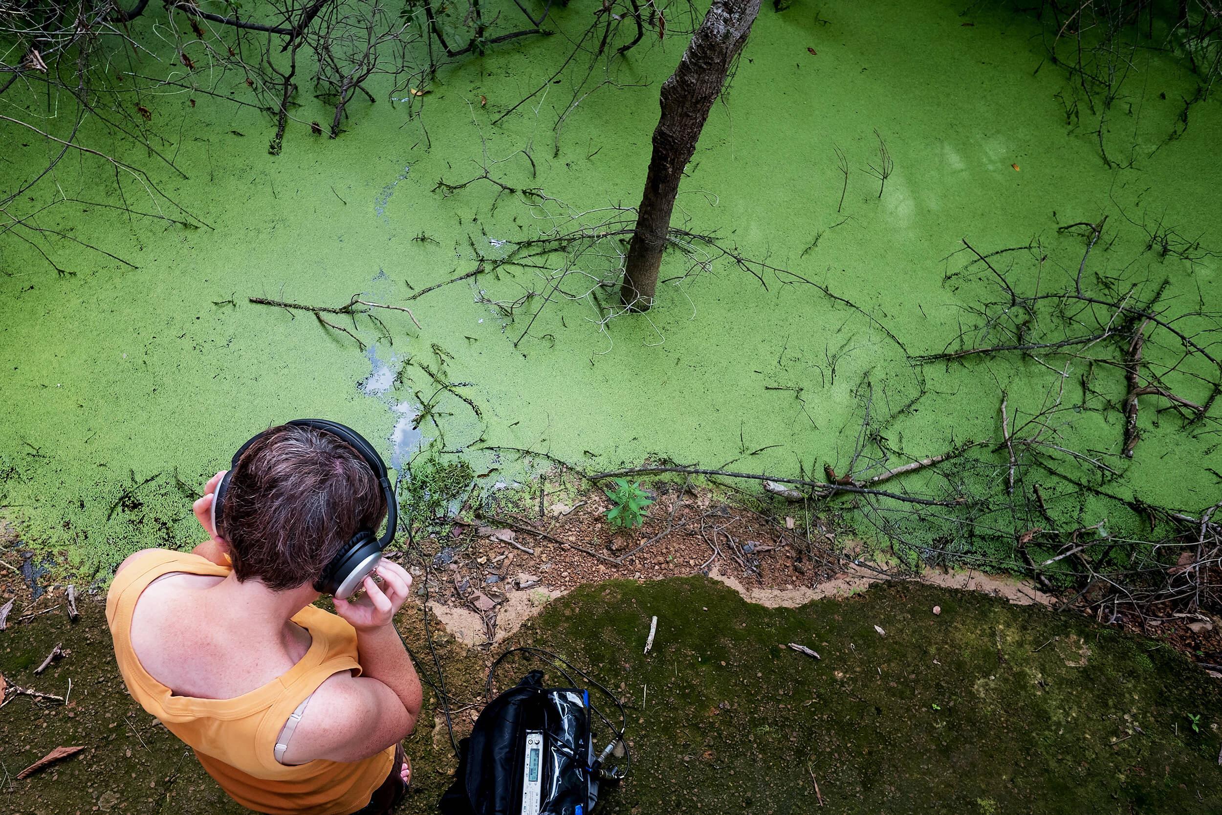 Karen Power listening to a recording device on headphones in front of a green body of water