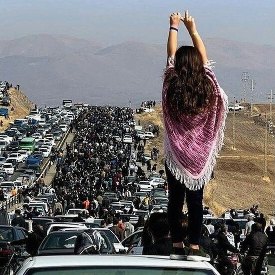 A woman stands on top of a car with her hair uncovered while a mass of people walk toward Jina Amini's grave in Saqqez, Iran.
