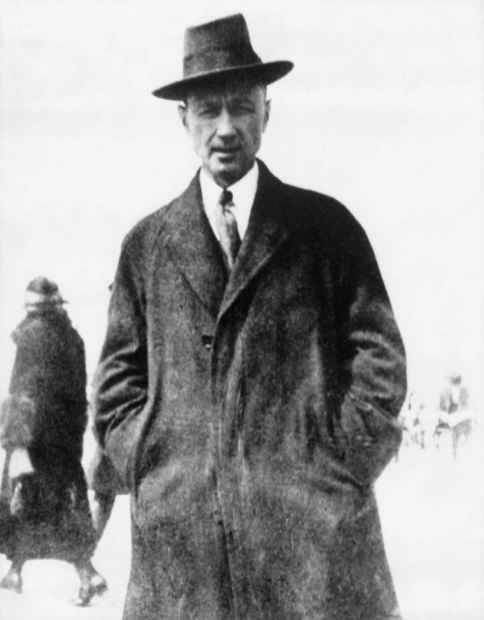 Charles Ives standing in a large coat with hands in pocket.