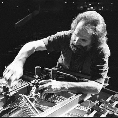 Trimpin bending over interior of piano, hands outstretched and making adjustments so as to modify the instrument.