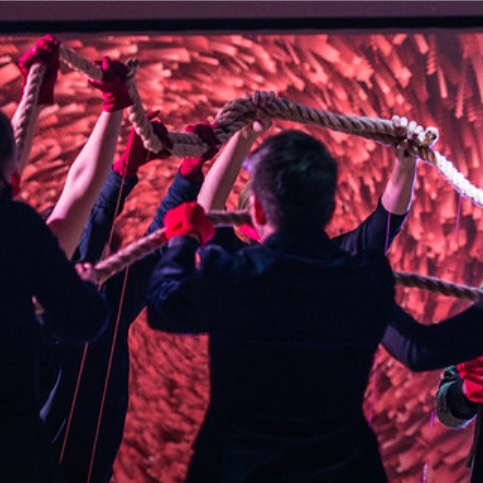 A group of people in dark clothing hold aloft a thick rope with strings attached to the rope and the floor.