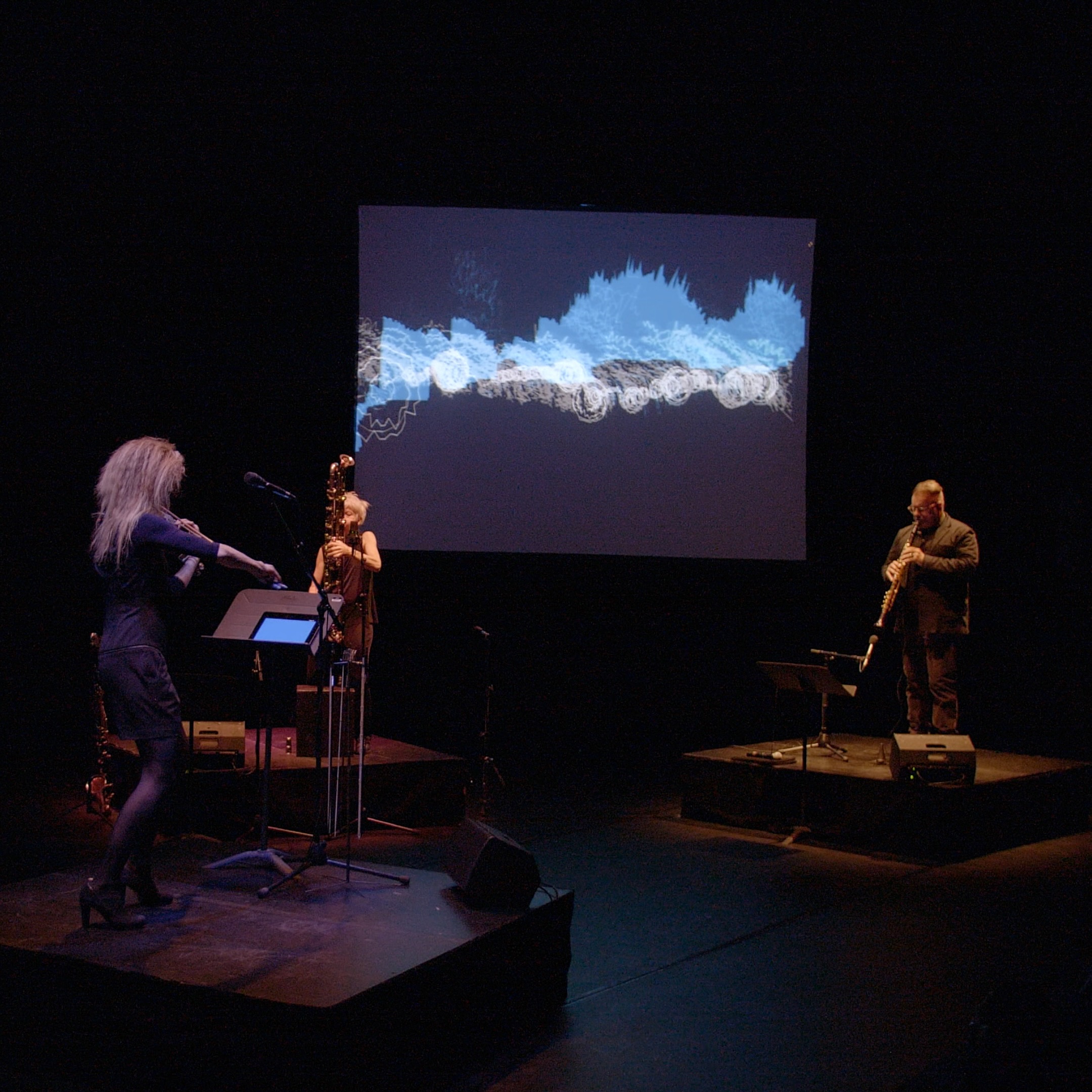 A trio performing in front of a screen with blue images.