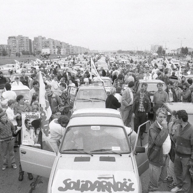 A line of cars surrounded by people. The hood of the first car reads "Solidarność"