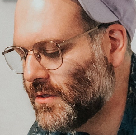 A handsome man with a beard, glasses, and a hat looking down and to the left.