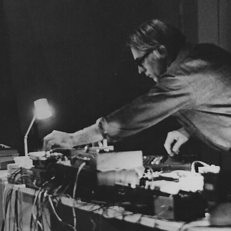 A black and white picture of a white man with glasses twisting knobs on musical devices.