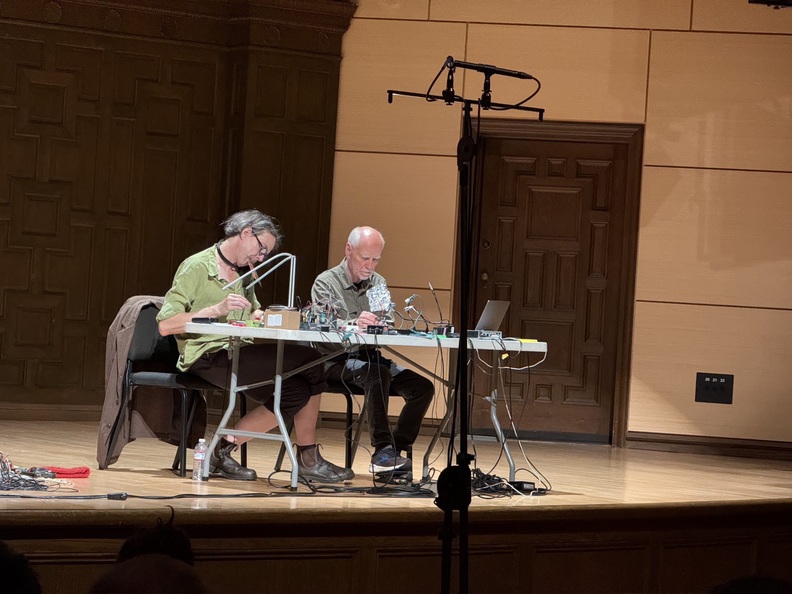 Two men sitting at a table playing sounds with objects.