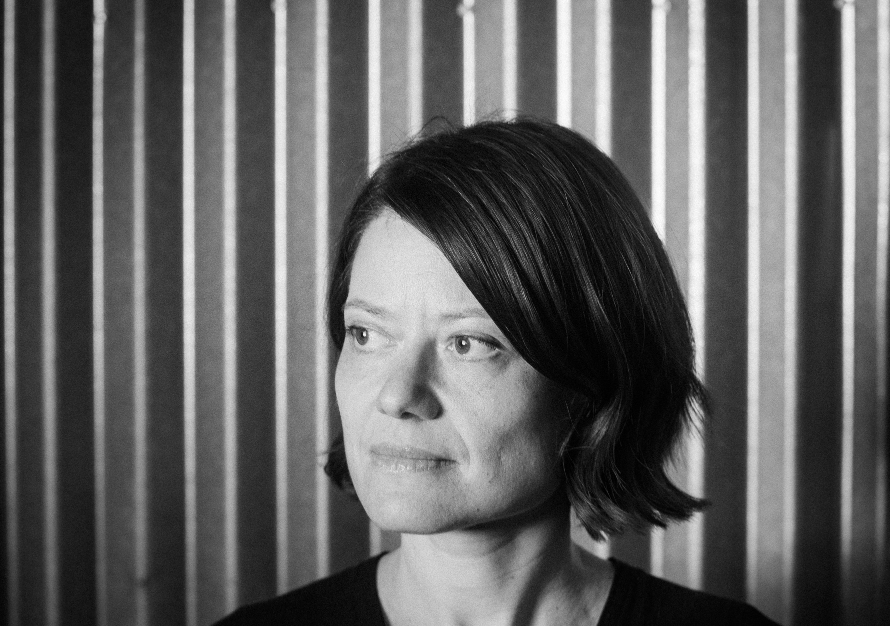 I woman looks to her right, audience left in front of a striped background.