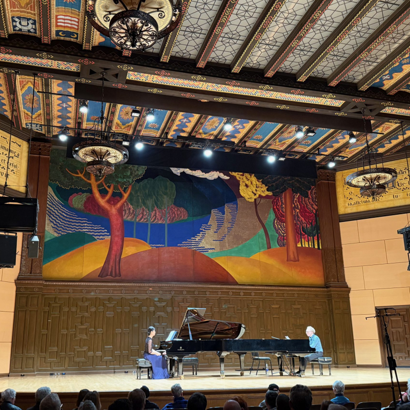 Gloria Cheng and Ralph van Raat performing on a piano in a large concert hall with a mural.