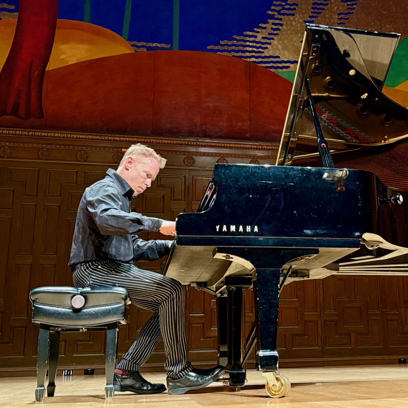 Geoffrey Burleson playing a grand piano on stage with a mural in the background.