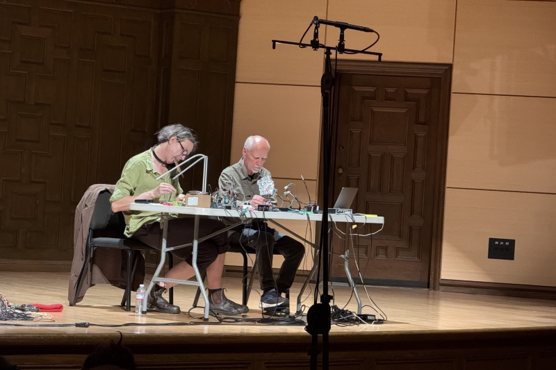 Two men sitting at a table playing sounds with objects.