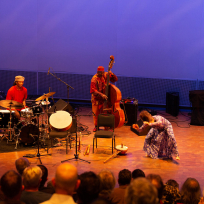 An ensemble with drumset, bass, and dancer.