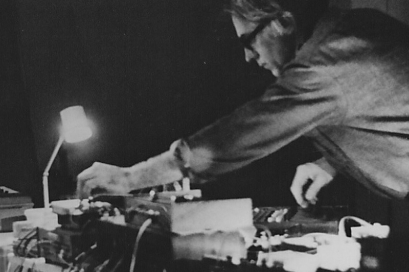 A black and white picture of a white man with glasses twisting knobs on musical devices.